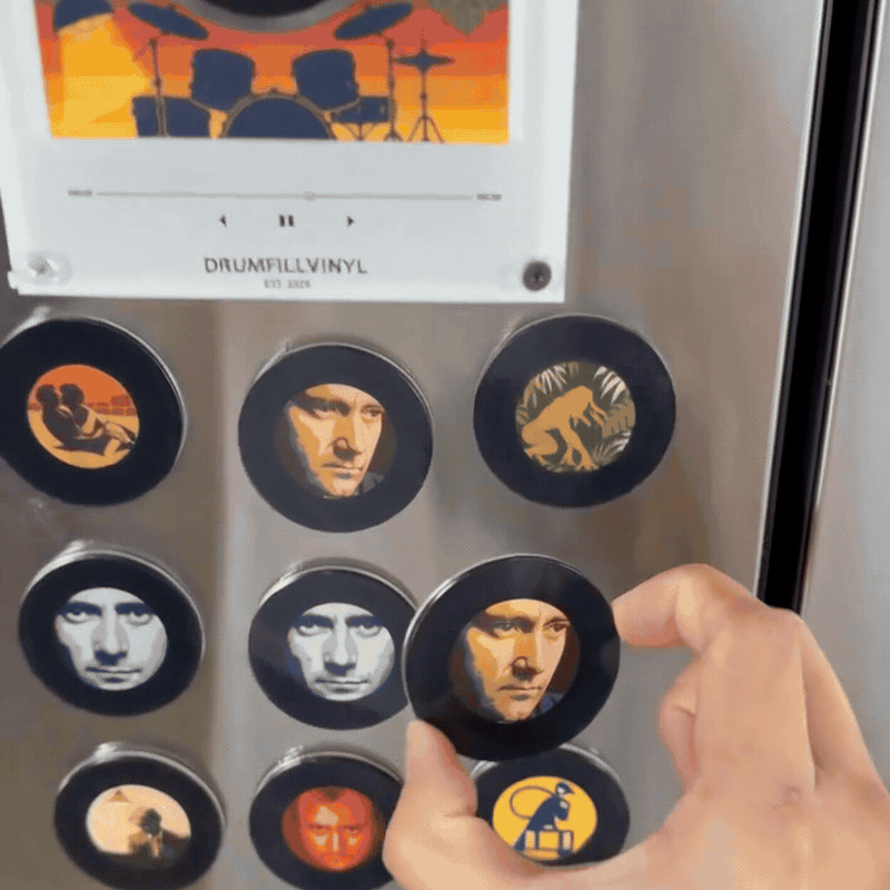 A hand lifts a mini vinyl disc off the fridge and drops it onto the spindle in the frame